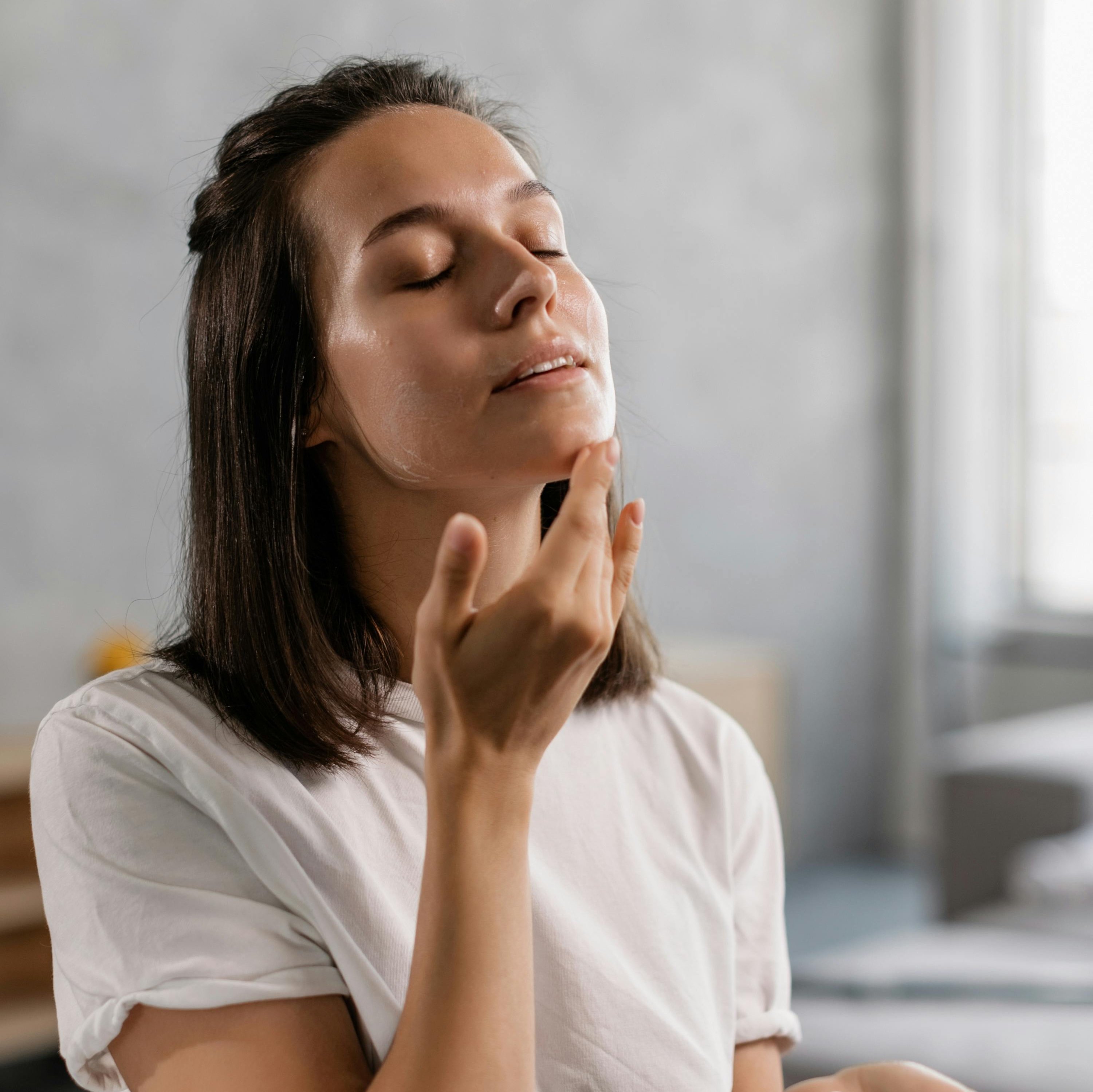 Frau mit geschlossenen Augen, die sanft Pflegeprodukt auf ihr Gesicht aufträgt, symbolisiert Hautpflege und Wohlbefinden im Kontext der Haarentfernung und Gesichtspflege.
