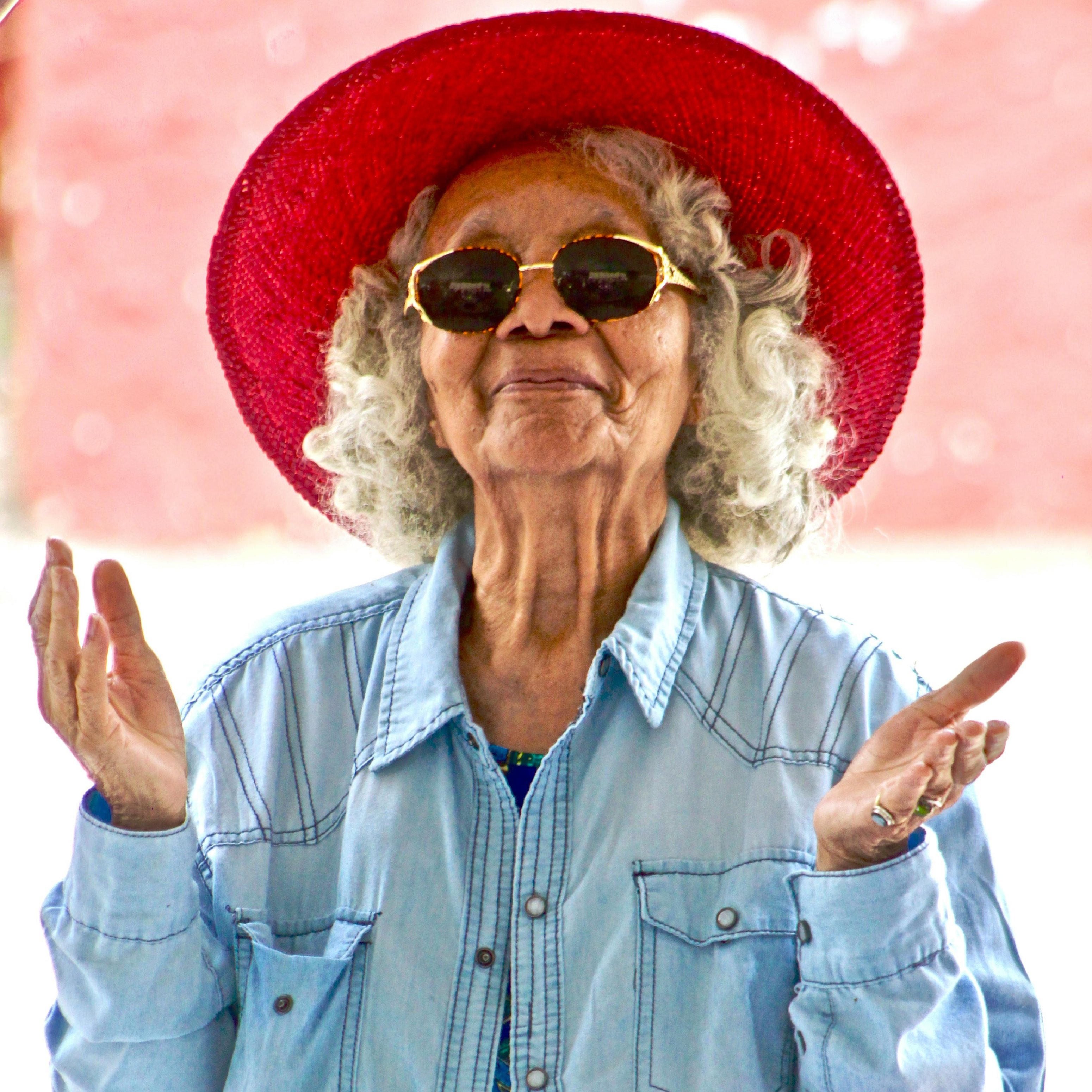 Ältere Frau mit lockigem, grauem Haar, rotem Hut und Sonnenbrille, die lächelnd die Hände hebt, umgeben von einem unscharfen, hellen Hintergrund, symbolisiert Freude und positive Ausstrahlung in einem Kontext von Hautpflege und Anti-Aging-Lösungen.