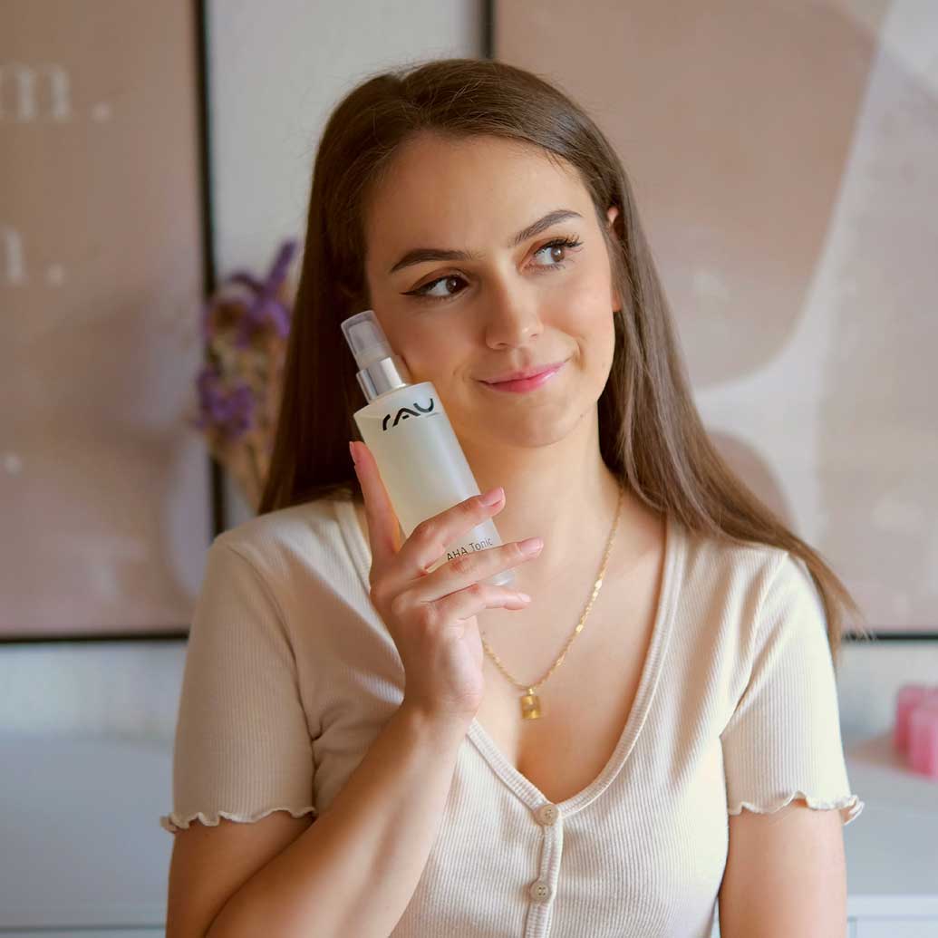 Frau hält das AHA-Tonic von RAU Cosmetics in der Hand, lächelnd, mit sanftem Hintergrund und dekorativen Elementen, betont die sanfte Exfoliation und Hautpflege.