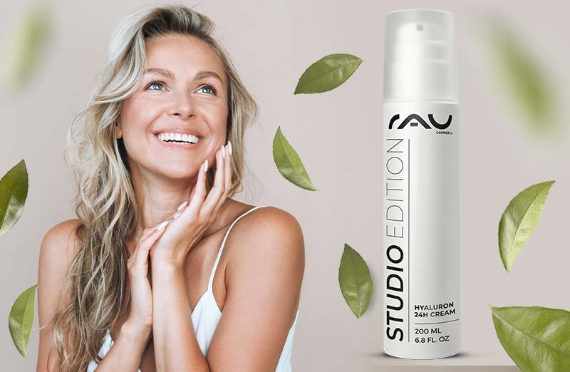 Frau mit leichtem Haar lächelt, präsentiert Hyaluron 24h Creme von RAU Cosmetics neben grünen Blättern.