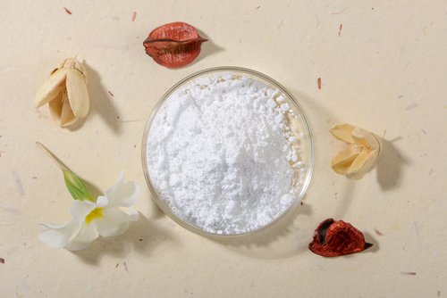 Pulverförmiger Allantoin in einer Glasschale, umgeben von getrockneten Blüten und Pflanzenmaterialien, symbolisiert die natürlichen Inhaltsstoffe in Hautpflegeprodukten.