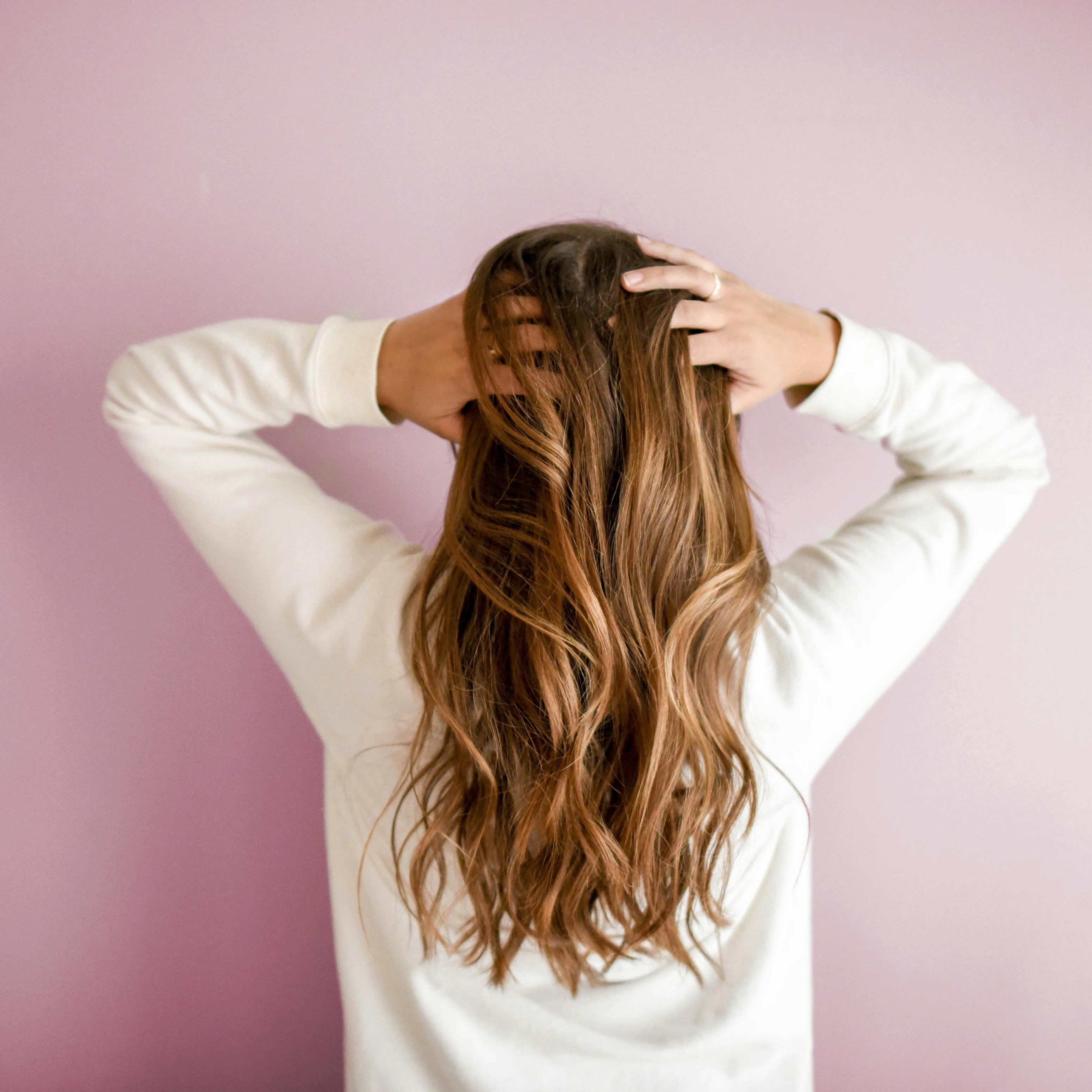 Frau mit langen, welligen Haaren, die sich am Kopf kratzt, vor einer rosa Wand, symbolisiert die Pflege von Kopfhaut und Haar, passend zu Niacinamide Serum für Schuppen und empfindliche Haut.