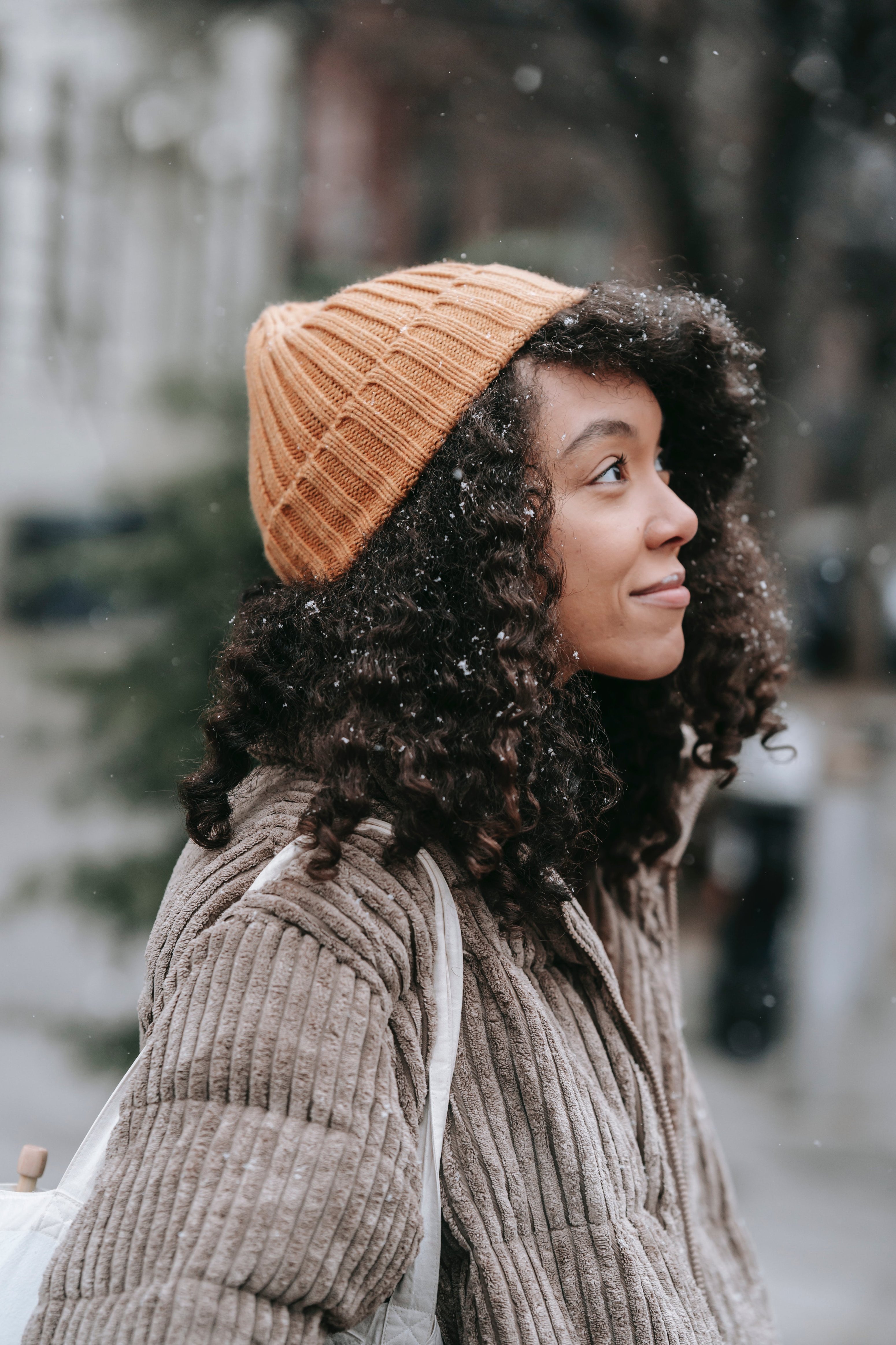 Frau mit lockigem Haar und orangefarbener Mütze, die in winterlicher Umgebung steht, symbolisiert die Bedeutung von Winterhautpflege mit RAU Cosmetics.