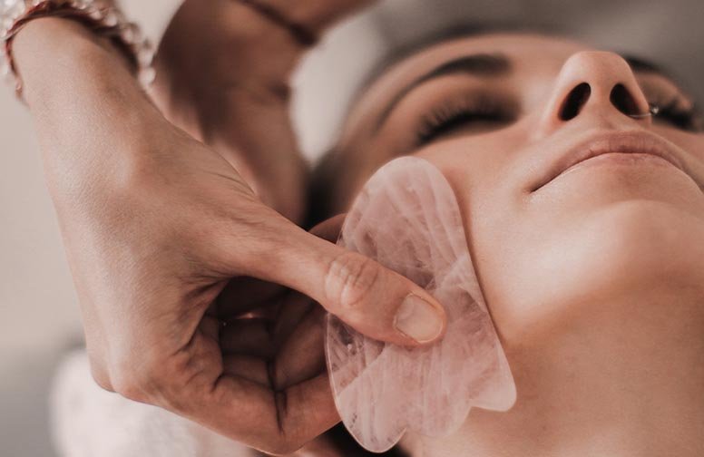 Gua Sha Gesichtsmassage mit Rosenquarzstein, Hand führt sanfte Massagebewegungen über das Gesicht einer Frau, Hautpflege und Entspannung.