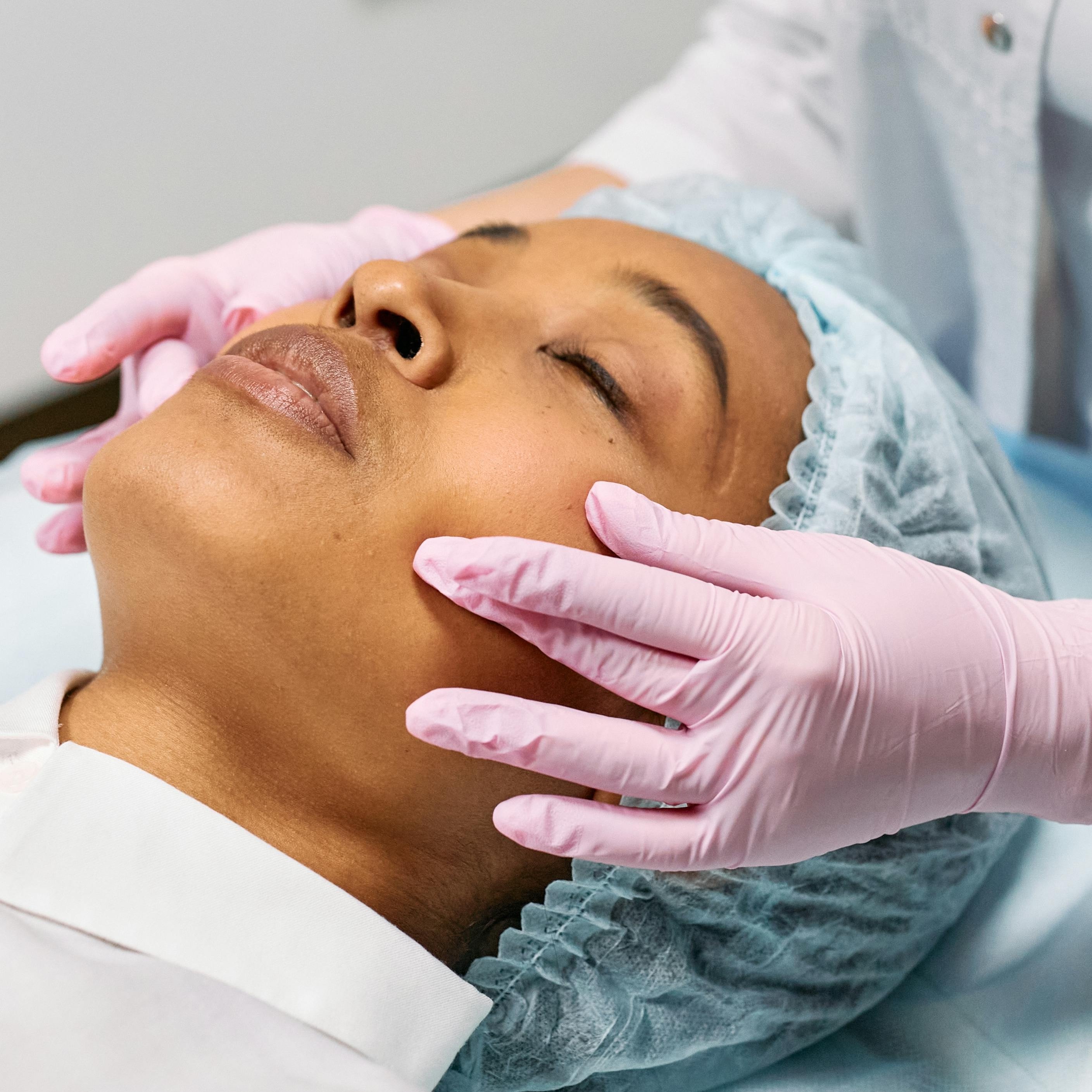 Frau mit geschlossenen Augen während einer Gesichtsbehandlung, Hände mit rosa Handschuhen massieren sanft das Gesicht, Fokus auf Hautpflege und ästhetische Behandlung.